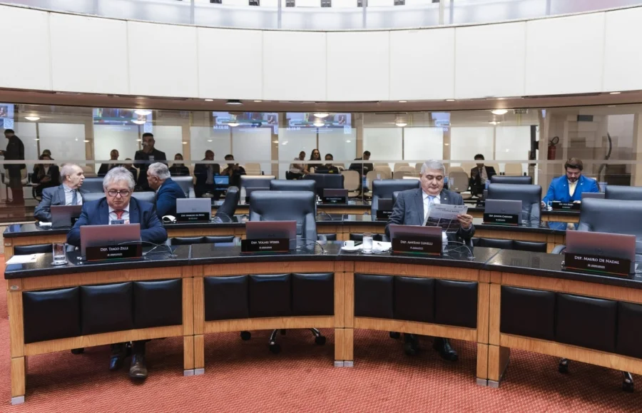 Deputados cobram rigor em sabatina de indicado ao STF Uma fotografia de uma sessão plenária na Assembleia Legislativa de Santa Catarina (Alesc). Deputados estaduais de Santa Catarina, incluindo o Deputado Mauro de Nadal (em terno azul ao fundo) e o Deputado Tiago Zeilli (segundo da esquerda), estão sentados em bancadas semicirculares e acolchoadas. Um deputado no centro lê um jornal (