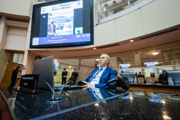 Deputados debatem segurança e saúde na Alesc Deputado Maurício Eskudlark sentado na bancada da Alesc durante sessão plenária em Florianópolis. Ao fundo, um telão exibe homenagem ao Dia do Policial Civil e Militar em Santa Catarina.