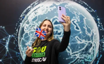Doutoranda da Unesc pesquisa teste de sífilis na Inglaterra Pesquisadora Amanda Christine da Silva Kursancew, da Unesc, segura uma pequena bandeira do Reino Unido e tira uma selfie em frente a um painel de mapa-múndi digital, celebrando sua partida para o doutorado sanduíche na Inglaterra.