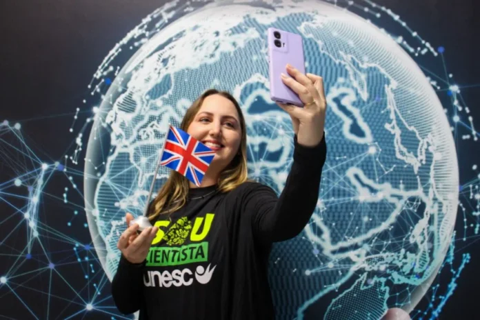 Doutoranda da Unesc pesquisa teste de sífilis na Inglaterra Pesquisadora Amanda Christine da Silva Kursancew, da Unesc, segura uma pequena bandeira do Reino Unido e tira uma selfie em frente a um painel de mapa-múndi digital, celebrando sua partida para o doutorado sanduíche na Inglaterra.
