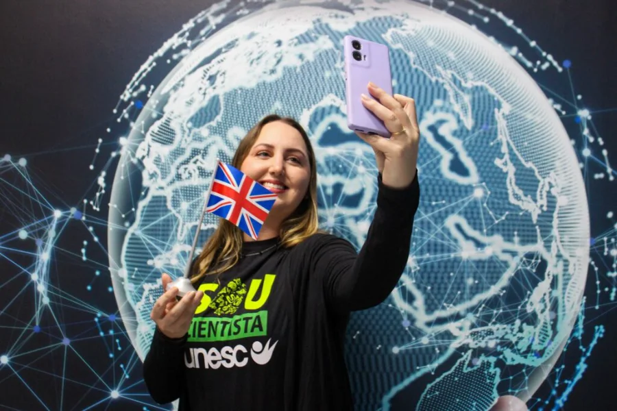 Pesquisadora Amanda Christine da Silva Kursancew, da Unesc, segura uma pequena bandeira do Reino Unido e tira uma selfie em frente a um painel de mapa-múndi digital, celebrando sua partida para o doutorado sanduíche na Inglaterra.