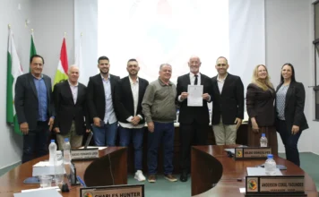 Edésio Loch assume vaga na Câmara de Forquilhinha Vereador Edésio Loch em pé, segurando seu diploma de posse ao lado de outros parlamentares no plenário da Câmara de Vereadores de Forquilhinha durante a 9ª Sessão Ordinária. A imagem mostra o grupo em frente às bandeiras e mesas com identificações dos vereadores Charles Hunter e Anderson Coral Fagundes.