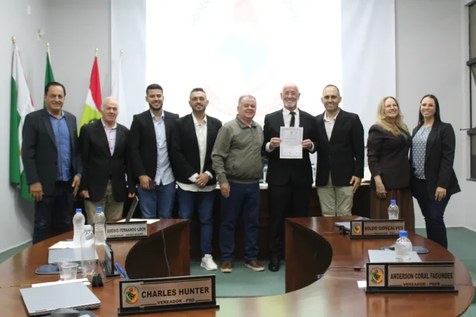 Edésio Loch assume vaga na Câmara de Forquilhinha Vereador Edésio Loch em pé, segurando seu diploma de posse ao lado de outros parlamentares no plenário da Câmara de Vereadores de Forquilhinha durante a 9ª Sessão Ordinária. A imagem mostra o grupo em frente às bandeiras e mesas com identificações dos vereadores Charles Hunter e Anderson Coral Fagundes.