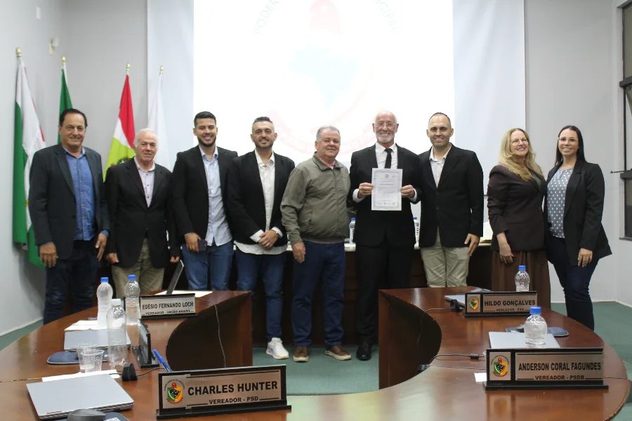 Vereador Edésio Loch em pé, segurando seu diploma de posse ao lado de outros parlamentares no plenário da Câmara de Vereadores de Forquilhinha durante a 9ª Sessão Ordinária. A imagem mostra o grupo em frente às bandeiras e mesas com identificações dos vereadores Charles Hunter e Anderson Coral Fagundes.
