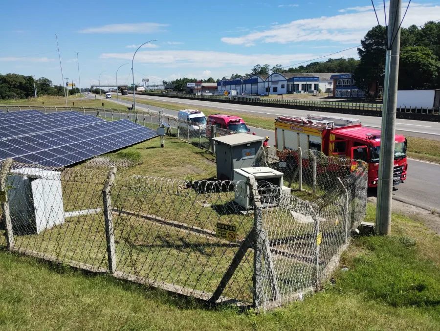 Caminhão do Corpo de Bombeiros e ambulância estacionados ao lado de usina de painéis solares atingida por incêndio e vandalismo em Criciúma, às margens de uma rodovia.