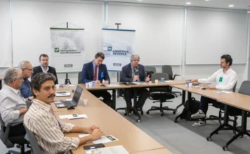 FIESC debate economia circular para plásticos de uso único Reunião estratégica na FIESC em Florianópolis, com Elias Caetano, diretor executivo do SINPLASC, falando ao microfone para apresentar o Projeto Defesa Circular sobre economia circular de plásticos de uso único. Ao fundo, banners do Comitê Estratégico para Logística Reversa e da Câmara de Meio Ambiente e Sustentabilidade são visíveis. Homens em ternos e roupas de trabalho estão sentados à mesa de conferência com laptops e materiais de apresentação.