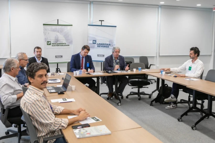 FIESC debate economia circular para plásticos de uso único Reunião estratégica na FIESC em Florianópolis, com Elias Caetano, diretor executivo do SINPLASC, falando ao microfone para apresentar o Projeto Defesa Circular sobre economia circular de plásticos de uso único. Ao fundo, banners do Comitê Estratégico para Logística Reversa e da Câmara de Meio Ambiente e Sustentabilidade são visíveis. Homens em ternos e roupas de trabalho estão sentados à mesa de conferência com laptops e materiais de apresentação.