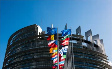 FIESC debate impactos do acordo Mercosul-União Europeia Fachada curva do prédio do Parlamento Europeu sob céu azul, com fileiras de bandeiras dos países membros da União Europeia em destaque, representando a governança e a unidade europeia.
