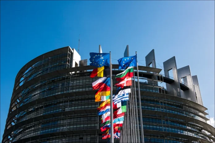 FIESC debate impactos do acordo Mercosul-União Europeia Fachada curva do prédio do Parlamento Europeu sob céu azul, com fileiras de bandeiras dos países membros da União Europeia em destaque, representando a governança e a unidade europeia.
