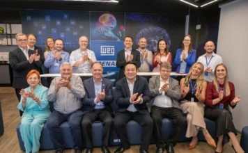 FIESC e WEG ampliam cooperação em ensino profissional Lideranças da FIESC, WEG e SENAI posam juntas e batem palmas em um centro de formação em Jaraguá do Sul, Santa Catarina, celebrando parceria e investimentos. Os logos da WEG e SENAI são visíveis no painel de fundo tecnológico.