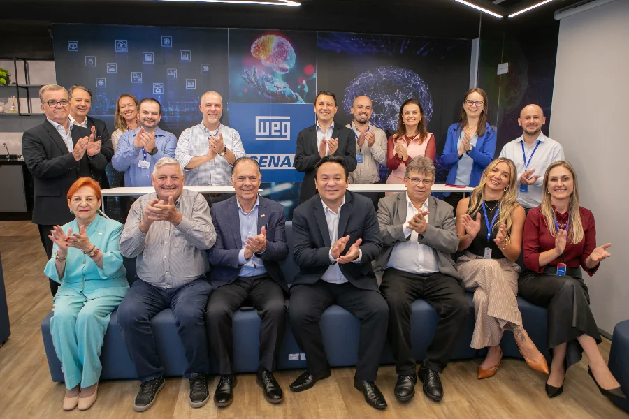 Lideranças da FIESC, WEG e SENAI posam juntas e batem palmas em um centro de formação em Jaraguá do Sul, Santa Catarina, celebrando parceria e investimentos. Os logos da WEG e SENAI são visíveis no painel de fundo tecnológico.