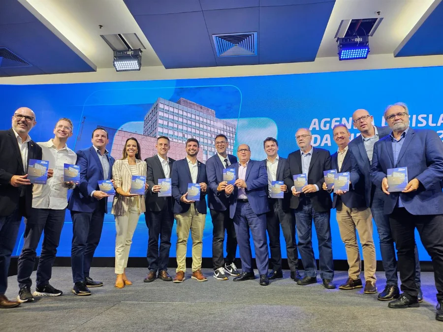 Fiergs entrega Agenda Legislativa 2026 à Assembleia Deputado Sergio Peres, Claudio Bier e parlamentares gaúchos posam segurando exemplares da Agenda Legislativa 2026 da Fiergs durante entrega oficial em Porto Alegre. O grupo está em um palco à frente de um telão azul que exibe a sede da federação.