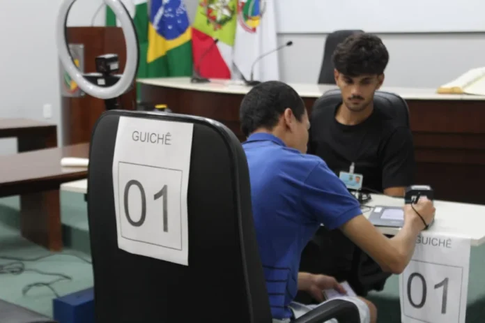 Forquilhinha registra 257 atendimentos em ação eleitoral Atendimento eleitoral sendo realizado em guichê na Câmara de Vereadores de Forquilhinha, com cidadão utilizando dispositivo eletrônico de biometria e assinatura para regularização de título de eleitor.