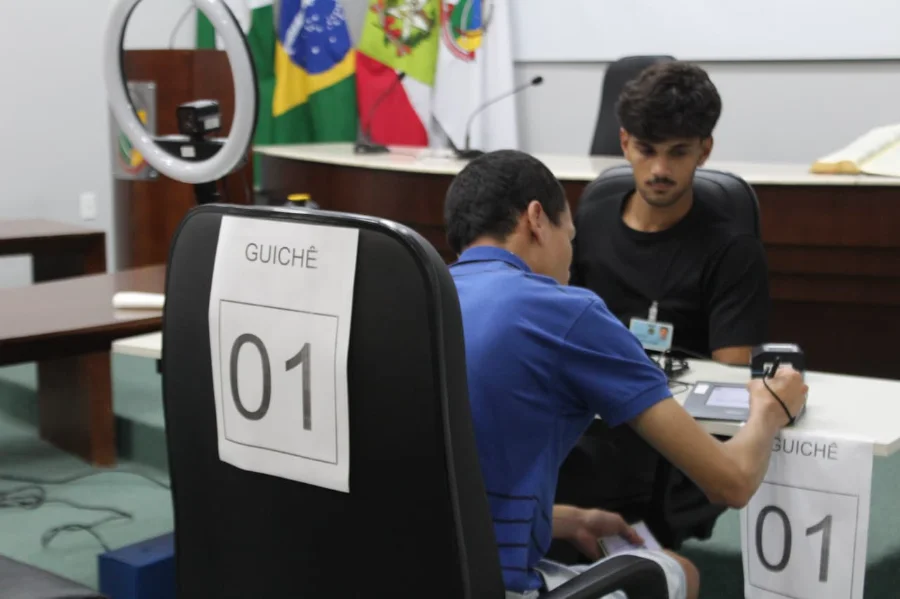 Atendimento eleitoral sendo realizado em guichê na Câmara de Vereadores de Forquilhinha, com cidadão utilizando dispositivo eletrônico de biometria e assinatura para regularização de título de eleitor.