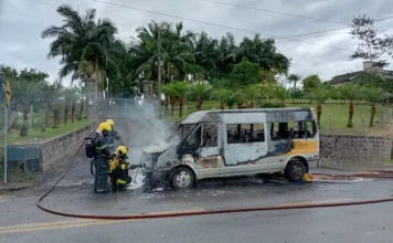 Furgão escolar pega fogo na rodovia SC-445 em Urussanga Três bombeiros de Urussanga (SC) em ação para extinguir o fogo que consome um furgão de transporte escolar na rodovia SC-445. O veículo está visivelmente carbonizado e há fumaça saindo do compartimento do motor.