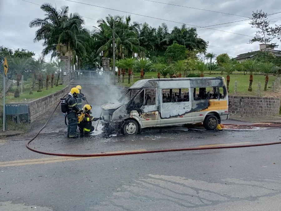 Três bombeiros de Urussanga (SC) em ação para extinguir o fogo que consome um furgão de transporte escolar na rodovia SC-445. O veículo está visivelmente carbonizado e há fumaça saindo do compartimento do motor.