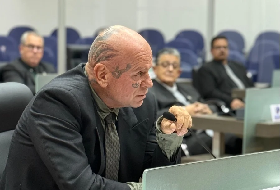 Vereador Jorge Ghiraldo, com tatuagens faciais visíveis, falando em um microfone durante uma sessão na Câmara de Vereadores sobre o aumento da violência contra a mulher. Ele veste um terno e gesticula.