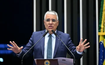 Girão defende impeachment de ministros do STF no Senado O senador Eduardo Girão (Novo-CE), de terno azul e óculos, faz um pronunciamento no púlpito do Plenário do Senado Federal. Ele está com as mãos levantadas e as palmas voltadas para a frente em um gesto de discurso, falando ao microfone. O púlpito exibe o brasão da República Federativa do Brasil, e ao fundo, à direita, vê-se a bandeira do Brasil. A foto captura o momento do seu discurso crítico ao STF e em defesa das prerrogativas do Senado.