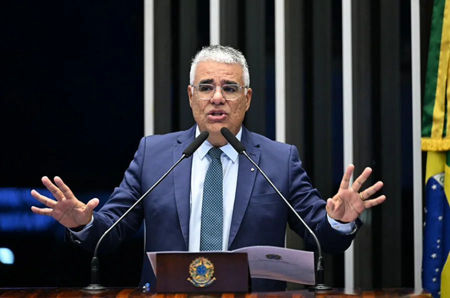 O senador Eduardo Girão (Novo-CE), de terno azul e óculos, faz um pronunciamento no púlpito do Plenário do Senado Federal. Ele está com as mãos levantadas e as palmas voltadas para a frente em um gesto de discurso, falando ao microfone. O púlpito exibe o brasão da República Federativa do Brasil, e ao fundo, à direita, vê-se a bandeira do Brasil. A foto captura o momento do seu discurso crítico ao STF e em defesa das prerrogativas do Senado.