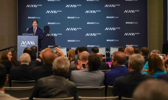 Governo estuda kits de TV 3.0 para famílias de baixa renda Ministro das Comunicações Frederico de Siqueira Filho discursa no NAB Show em Las Vegas sobre a implementação da TV 3.0 e a distribuição de kits para famílias de baixa renda. A imagem mostra o ministro ao púlpito com a logomarca do evento ao fundo e a plateia acompanhando o anúncio.