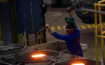 Governo libera R$ 15 bilhões para setores afetados por crises Indústria siderúrgica: trabalhador de macacão azul e capacete de proteção operando haste para remover impurezas de metal fundido incandescente em caldeira industrial. Foco em processo de fundição e segurança do trabalho.