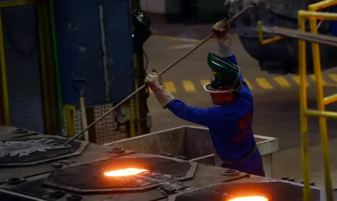 Governo libera R$ 15 bilhões para setores afetados por crises Indústria siderúrgica: trabalhador de macacão azul e capacete de proteção operando haste para remover impurezas de metal fundido incandescente em caldeira industrial. Foco em processo de fundição e segurança do trabalho.