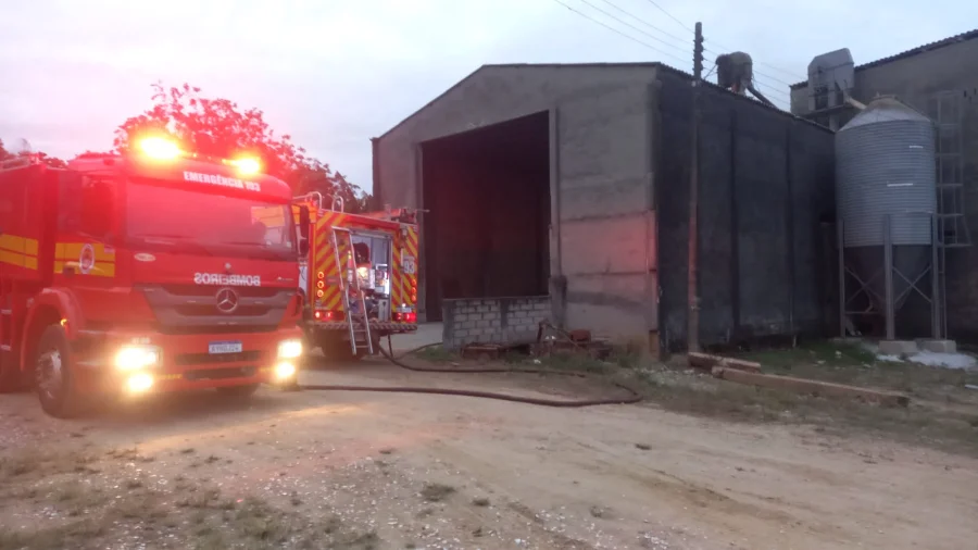 Caminhão de bombeiros (ABTR-208 do CBMSC) com luzes de emergência acesas em frente a um silo de grãos e uma edificação industrial de concreto em Içara, SC. Mangueiras de incêndio no chão indicam o combate a incêndio no local no bairro Vila Nova.