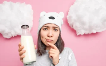 Intolerância à lactose ou alergia ao leite: você sabe a diferença? Mulher jovem com expressão pensativa segura uma garrafa de leite em um cenário rosa com nuvens falsas. A foto ilustra a confusão entre os sintomas da intolerância à lactose e a alergia ao leite (APLV).