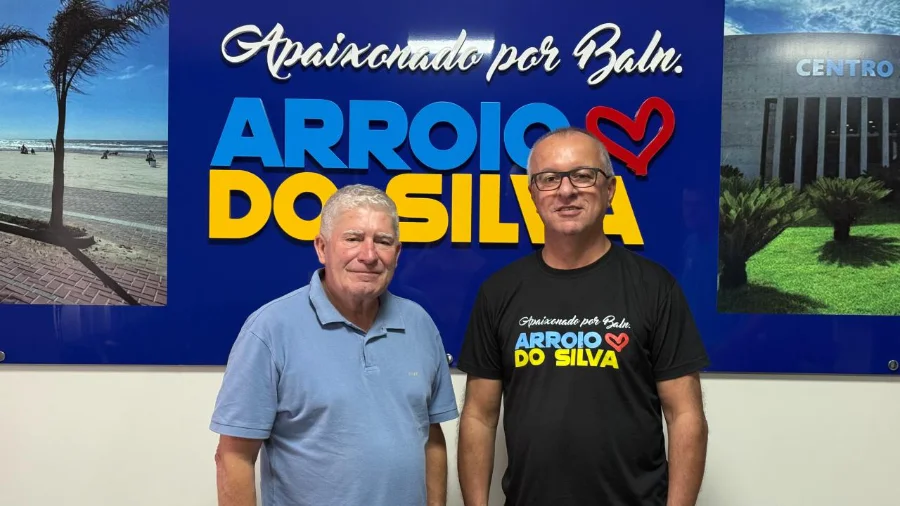 O prefeito Jorge Luiz Freitas e o novo secretário de Administração e Finanças, Amarante Jairo de Matos, posam juntos em frente ao painel institucional da Prefeitura de Balneário Arroio do Silva.