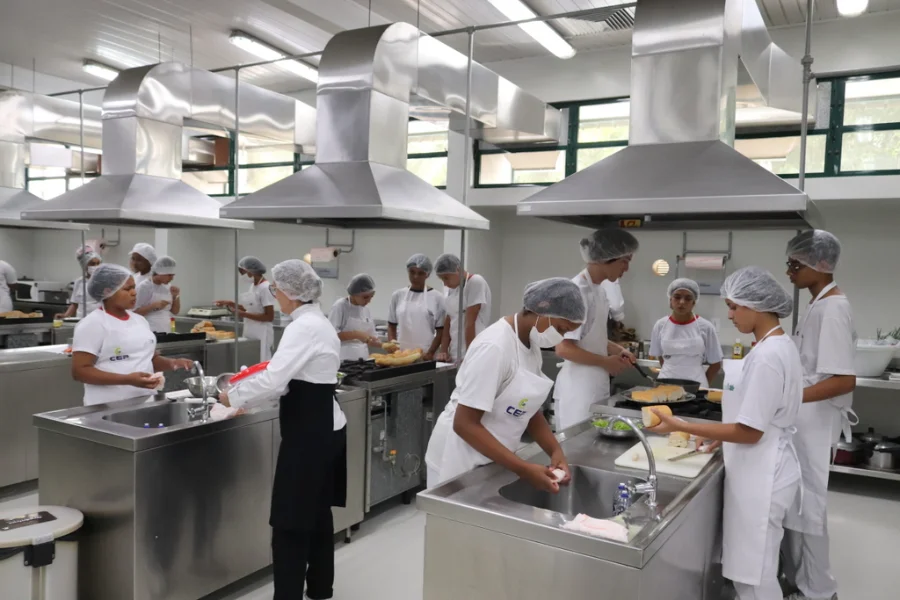Estudantes do Bairro da Juventude em aula prática de gastronomia no laboratório da Unesc, aprendendo técnicas de manipulação de alimentos e preparo de sanduíches em bancadas de aço inox.