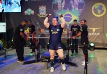 Legiane Civiero vence Arnold Classic South American 2026 Atleta catarinense Legiane Civiero celebra vitória no Arnold Classic South American 2026. A imagem mostra a competidora de powerlifting com os braços levantados e expressão de vibração em frente ao banco de supino, vestindo o uniforme azul da delegação brasileira.