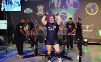 Legiane Civiero vence Arnold Classic South American 2026 Atleta catarinense Legiane Civiero celebra vitória no Arnold Classic South American 2026. A imagem mostra a competidora de powerlifting com os braços levantados e expressão de vibração em frente ao banco de supino, vestindo o uniforme azul da delegação brasileira.