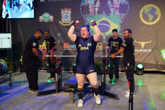 Legiane Civiero vence Arnold Classic South American 2026 Atleta catarinense Legiane Civiero celebra vitória no Arnold Classic South American 2026. A imagem mostra a competidora de powerlifting com os braços levantados e expressão de vibração em frente ao banco de supino, vestindo o uniforme azul da delegação brasileira.