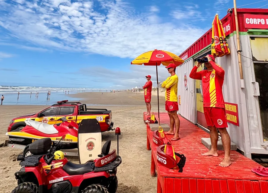 Três bombeiros guarda-vidas do CBMSC em vigilância na praia do litoral sul de Santa Catarina. O posto de salvamento exibe equipamentos de resgate como um jet ski e um quadriciclo vermelho com insígnias do Corpo de Bombeiros. A orla e banhistas ao fundo sob céu azul.
