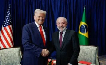 Lula repudia ataque contra Donald Trump em Washington Presidente Luiz Inácio Lula da Silva e Donald Trump apertam as mãos em um aperto de mão formal, posicionados entre as bandeiras do Brasil e dos Estados Unidos em um ambiente oficial em Washington.