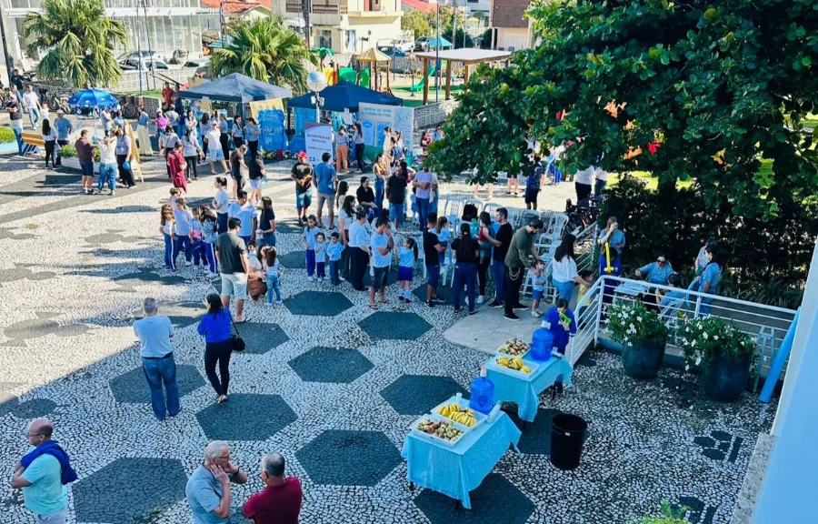 Vista aérea da praça central de Meleiro durante ação integrada da prefeitura, mostrando tendas de atendimento, profissionais de saúde e educação, e grande movimentação de moradores em um dia ensolarado.
