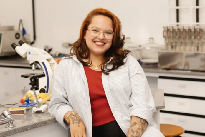 Mellanie Dutra palestra sobre ciência na Unesc nesta sexta Mellanie Fontes Dutra, pesquisadora e biomédica, sorrindo em um laboratório de pesquisa com um microscópio ao fundo, representando a divulgação científica na Unesc.