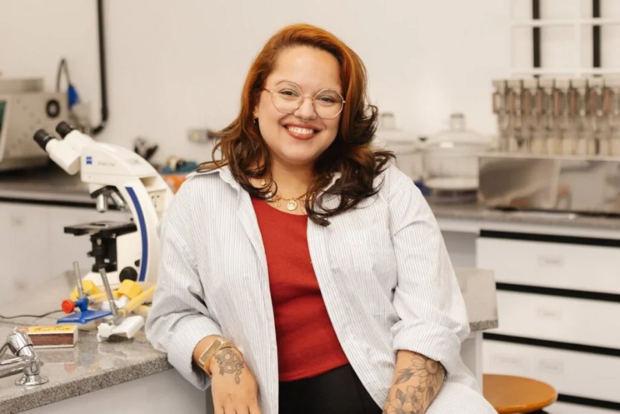 Mellanie Fontes Dutra, pesquisadora e biomédica, sorrindo em um laboratório de pesquisa com um microscópio ao fundo, representando a divulgação científica na Unesc.