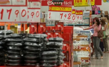 Mercado eleva previsão da inflação para 4,36% em 2026 Consumidora observa preços em prateleiras de loja de departamentos com etiquetas de ofertas em destaque. A imagem ilustra o setor de comércio e o impacto da inflação e do IPCA no poder de compra dos brasileiros.