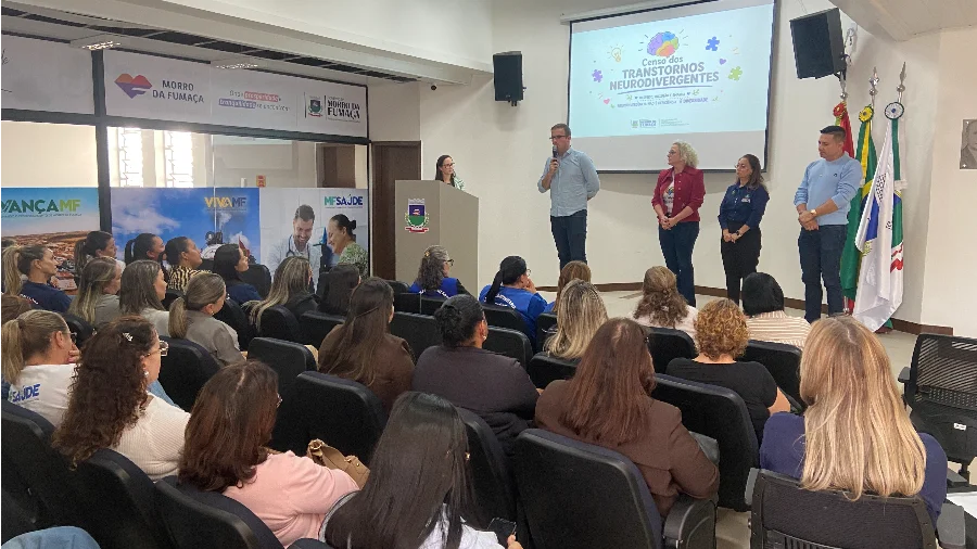 Prefeito Eduardo Sartor Guollo discursa ao microfone durante o lançamento do Censo dos Transtornos Neurodivergentes em Morro da Fumaça, ao lado de autoridades e diante de uma plateia no Salão de Atos.