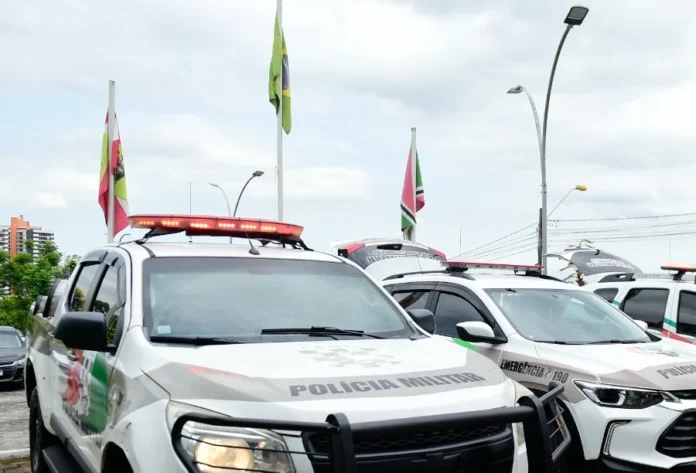 Motorista embriagado é preso após causar acidente em Criciúma Viaturas da Polícia Militar de Santa Catarina (PMSC) caracterizadas e estacionadas com o giroflex aceso, em frente a mastros com bandeiras, ilustrando operações policiais e a presença da força de segurança no estado.