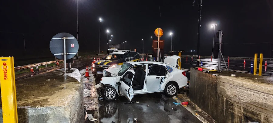 Acidente fatal na BR-101 em São João do Sul mostra veículo branco com a frente destruída após colisão frontal contra mureta de concreto em praça de pedágio durante a noite.