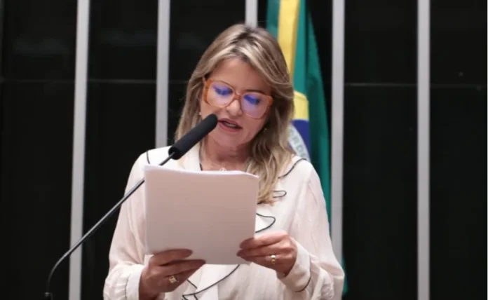 Novas regras contra violência financeira ao idoso avançam Deputada Flávia Morais (PDT-GO), relatora do projeto de lei, falando ao microfone enquanto segura e lê um documento oficial no plenário da Câmara dos Deputados. Ela usa óculos de armação laranja e uma blusa branca com detalhes pretos. A bandeira do Brasil é visível ao fundo.