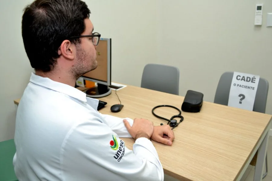 Profissional de saúde com jaleco da Unesc sentado em consultório diante de uma cadeira vazia com o cartaz 