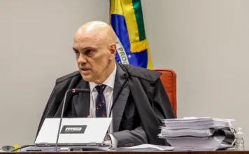 PGR e AGU barram tentativa do PT de limitar delações O ministro Alexandre de Moraes, do STF, sentado em sua bancada de trabalho com notebook e pilhas de processos, tendo ao fundo a bandeira do Brasil. A imagem ilustra o contexto jurídico da ação que busca limitar delações premiadas.