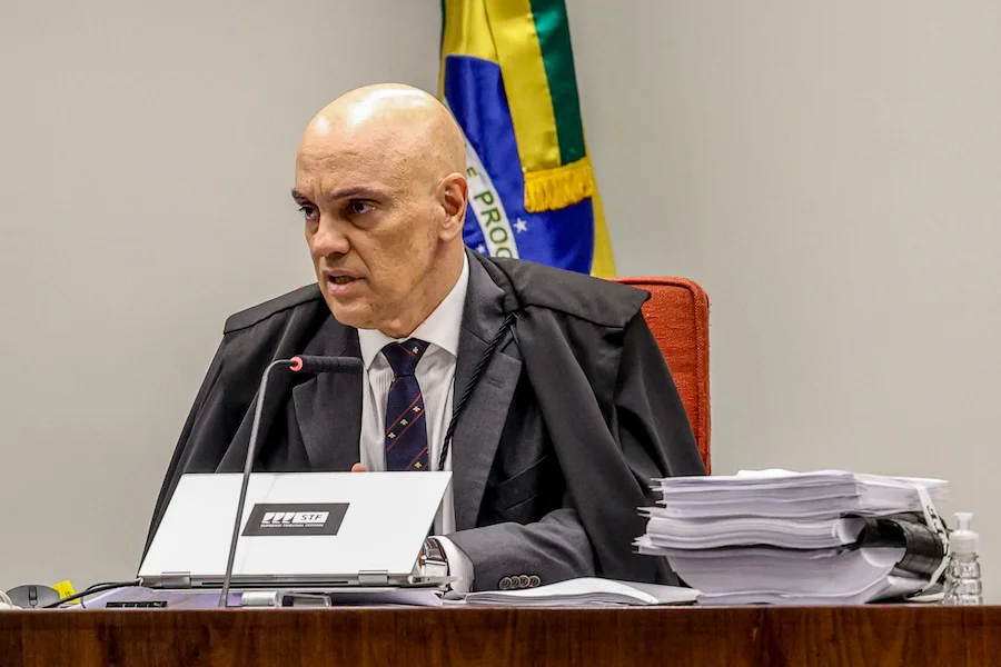 O ministro Alexandre de Moraes, do STF, sentado em sua bancada de trabalho com notebook e pilhas de processos, tendo ao fundo a bandeira do Brasil. A imagem ilustra o contexto jurídico da ação que busca limitar delações premiadas.