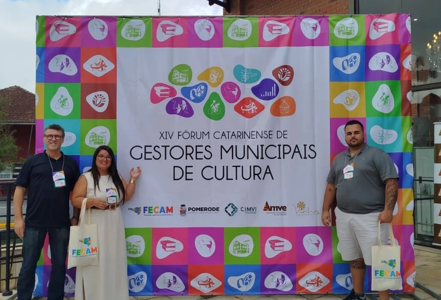 Secretária Alessandra da Rosa e o diretor Jaime Batista, de Passo de Torres, em frente ao banner do XIV Fórum Catarinense de Gestores Municipais de Cultura em Pomerode. Representantes participam de evento da FECAM sobre gestão cultural e economia criativa.