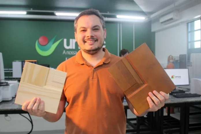 Pesquisa na Unesc transforma restos de madeira em lucro O pesquisador Mateus Alexandre Floriano sorri enquanto segura duas amostras de painéis de madeira de pinus reaproveitadas, em um laboratório da Unesc. A imagem ilustra o estudo de economia circular para o reaproveitamento de resíduos sólidos na indústria de portas do Sul catarinense.
