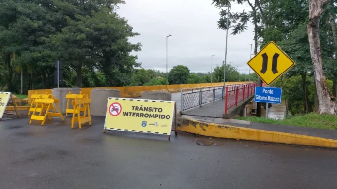 Ponte Giácomo Mazzuco será liberada para tráfego no sábado Entrada da Ponte Giácomo Mazzuco em Araranguá com placa amarela de trânsito interrompido e barreiras físicas. Ao fundo, a estrutura da ponte sobre o rio no bairro Barranca, sinalizando a interdição para obras de recuperação estrutural.