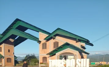 Pórtico de Timbé do Sul passa por revitalização completa Pórtico de entrada de Timbé do Sul com letreiro branco em destaque durante serviço de revitalização. A imagem mostra a estrutura arquitetônica com telhado verde, paredes de tijolos e uma colaboradora da Secretaria de Obras realizando a manutenção e pintura do local sob céu azul.